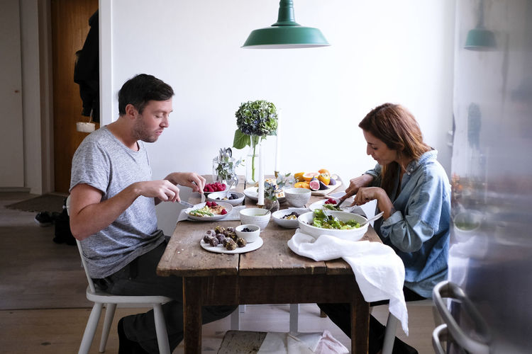 Side view of couple having food at table while sitting by wall