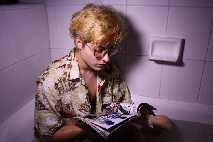 Teenage boy reading magazine while sitting in bathtub