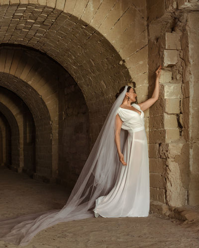 A sad elegant lady in white bride dress with a train languishes in the gloomy old castle