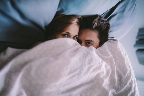 Portrait of young couple peeking over blanket lying on bed at home