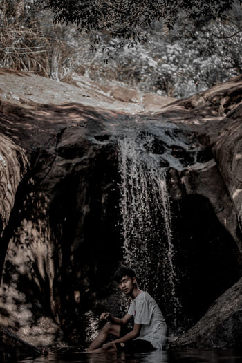 Full length of young man sitting by waterfall against cave