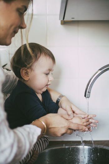 Mother helping baby girl washing hands under sink in kitchen