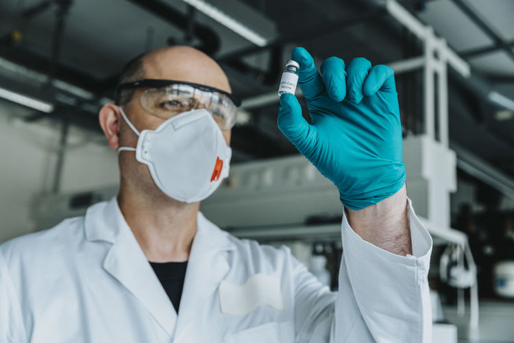 Scientist with protective face mask and eyeglasses holding vaccine bottle while standing at laboratory