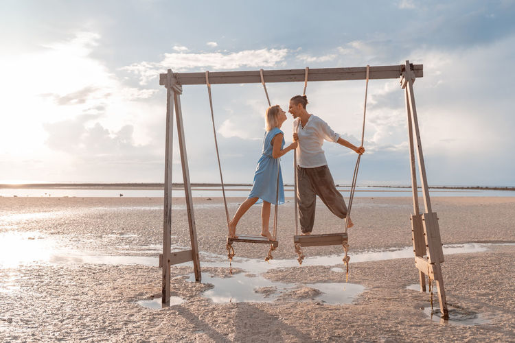 Man and a woman riding on a swing against the rising sun on the seashore