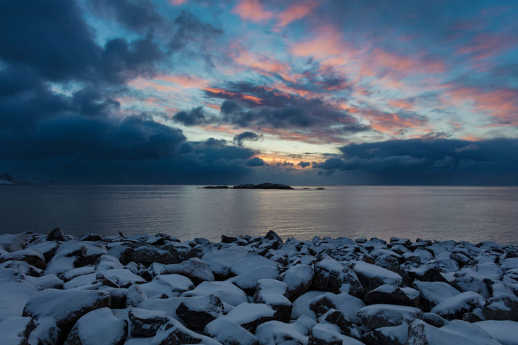 Snow covered rocky shore by sea against cloudy sky during sunset