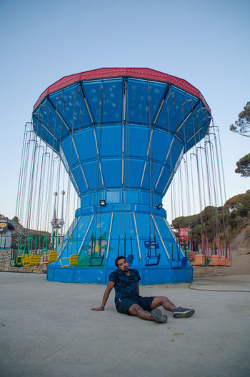 Man sitting in amusement park against clear blue sky