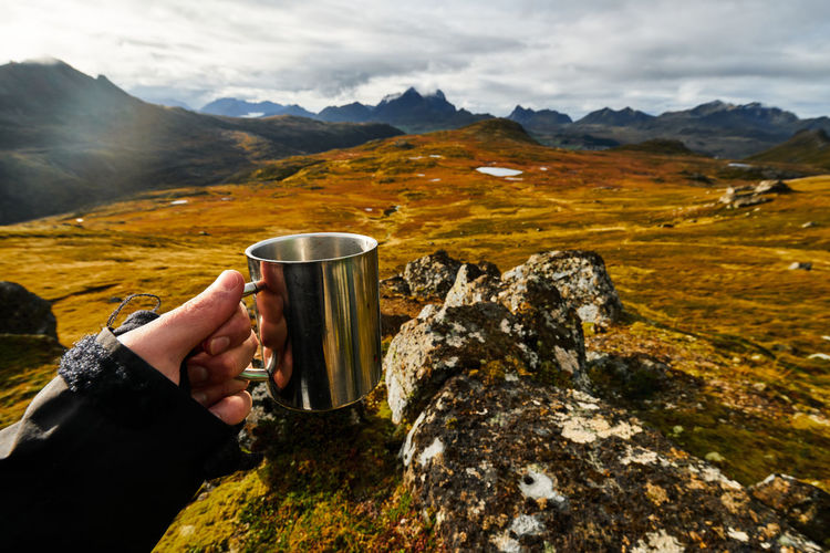 Hiker holding coffee cup with hand with mountains and landscape in the background on lofoten islands
