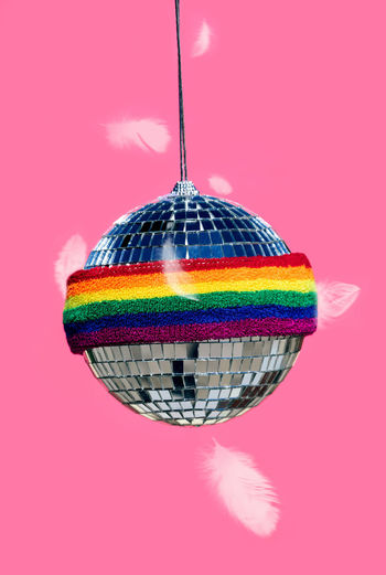 Scrunchy bright rainbow lgbt colors on small shining silver disco ball hanging on thread against pink background in light studio with feathers