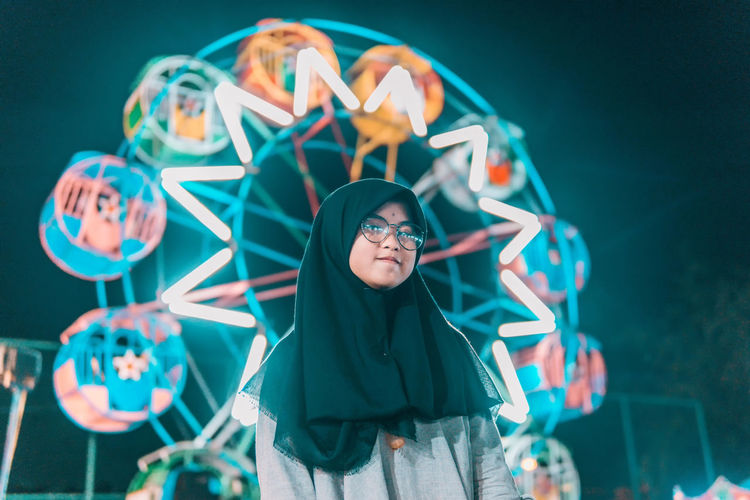 Portrait of young woman standing against illuminated ferris wheel at night