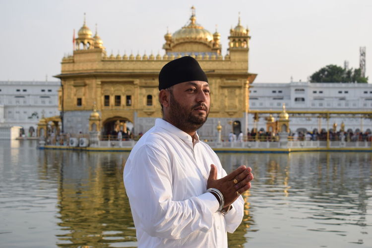 Man with hands clasped standing by pond against golden temple