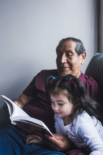 Senior ethnic grandfather reading interesting story in book for cute little girl while relaxing together on sofa at home
