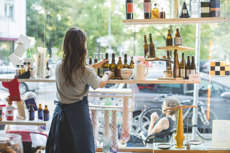 Rear view of female employee arranging bottles on shelf in deli