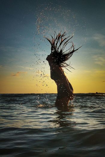 Side view of woman tossing hair in sea against sky during sunset