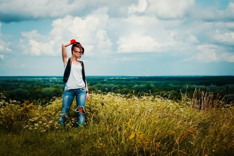 Hipster young woman with hand in hair standing on field against sky