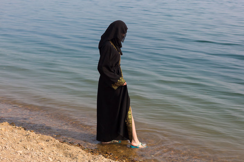 A muslim woman in national clothes wets her feet in sea.