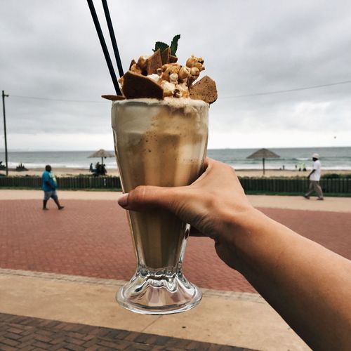 Cropped image of hand holding milkshake against sea and cloudy sky