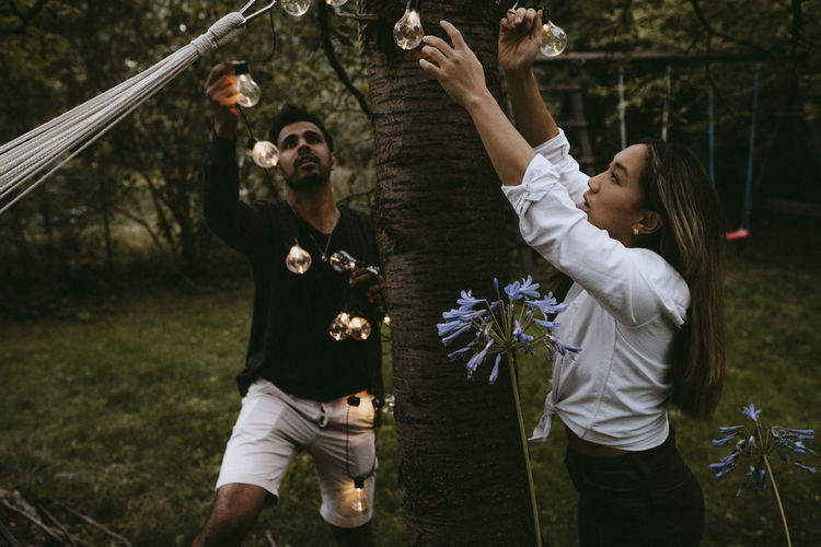 Male and female friends hanging lighting equipment in yard during dinner party