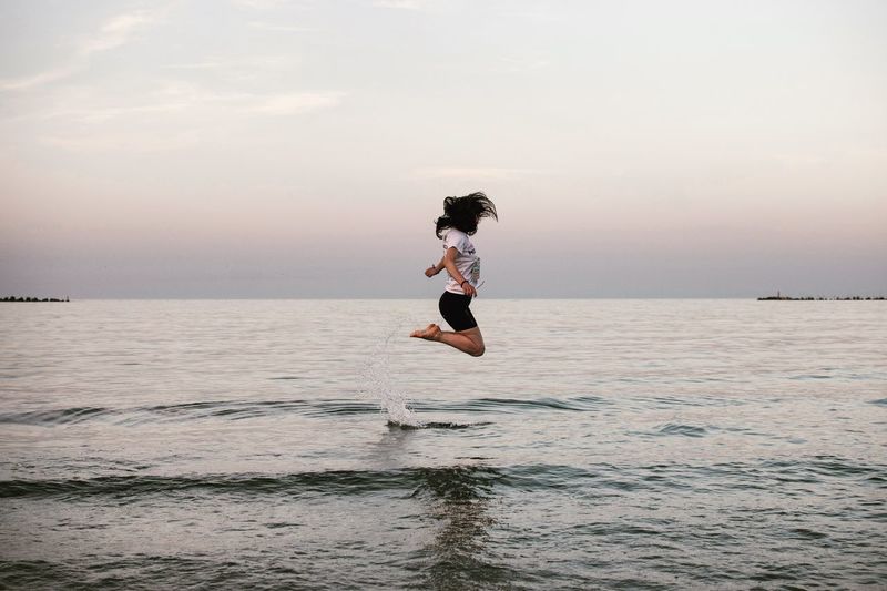 Woman jumping in sea against sky during sunset