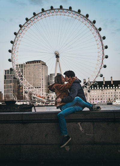 Side view of couple kissing while sitting against ferris wheel in city