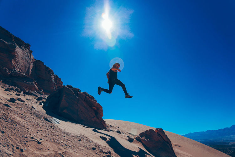 Low angle view of young man jumping on mountain against blue sky during sunny day