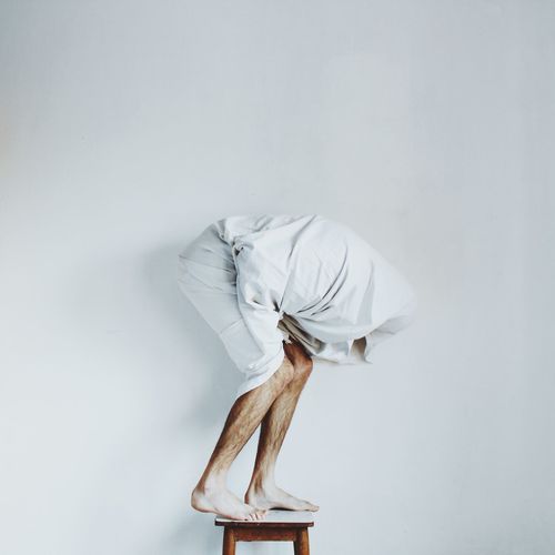 Man wrapped in blanket bending while standing on stool against white wall