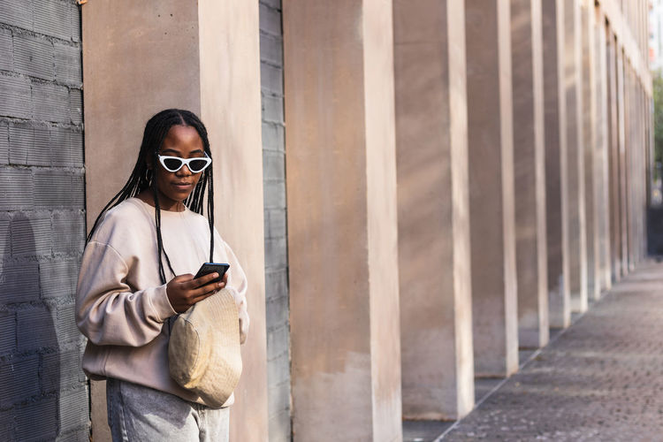 Young ethnic female in casual outfit leaning on gray brick wall and browsing smartphone on city street