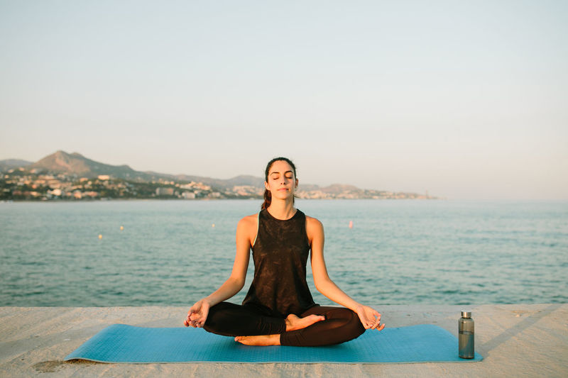 Relaxed female sitting on yoga mat in padmasana with mudra hands and practicing mindfulness with closed eyes on background of seascape at sunset