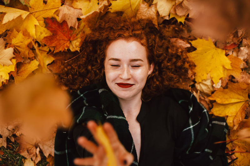 Happy redhead woman lying on field with orange dry leaves in autumn