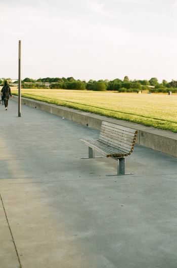 Empty bench on field by road against sky