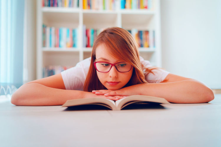 Beautiful woman wearing eyeglasses reading book lying on floor at home