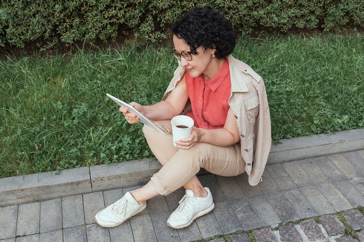 An adult woman sits on the lawn, drinks coffee and looks into a tablet.
