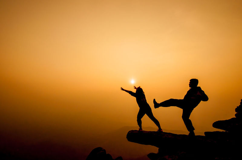 Silhouette man kicking woman from cliff against sky during sunset