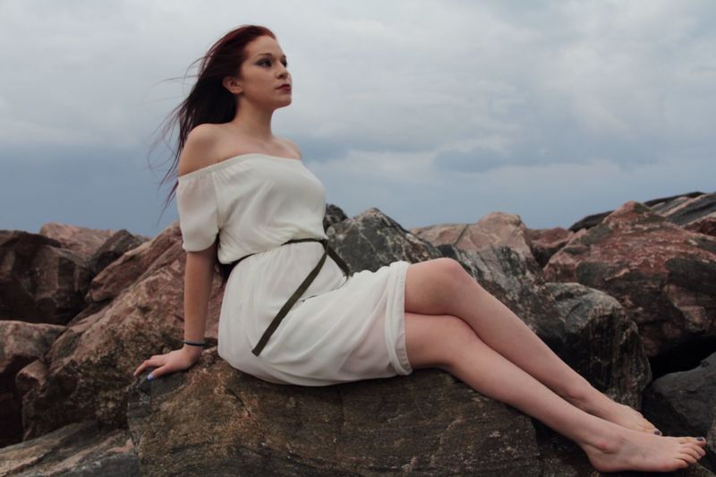Full length of thoughtful young woman looking away while sitting on rocks at beach against sky