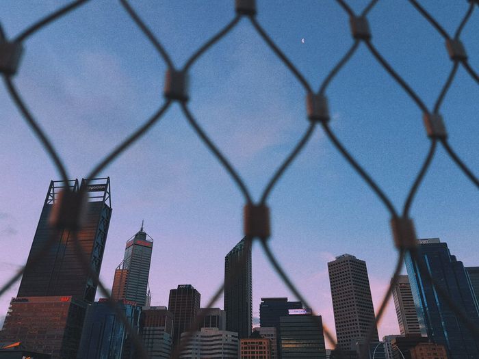 Low angle view of modern buildings against sky seen through chainlink fence