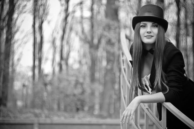 Portrait of young woman wearing hat standing by railing against trees