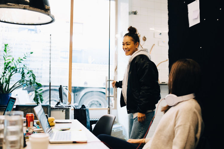 Smiling young businesswoman holding door while standing by female colleague at creative office