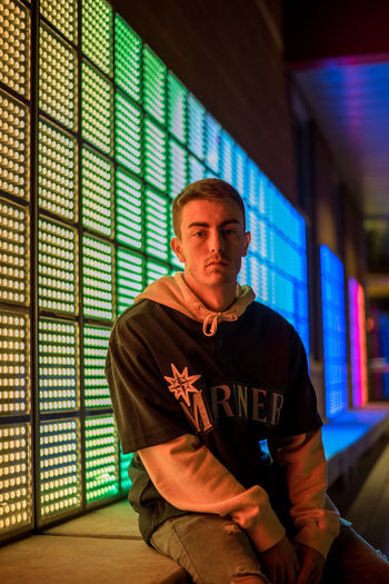 Portrait of serious young man sitting by illuminated building at night