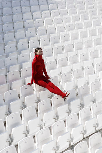 Full length of fashion model in red sitting on white chairs at stadium