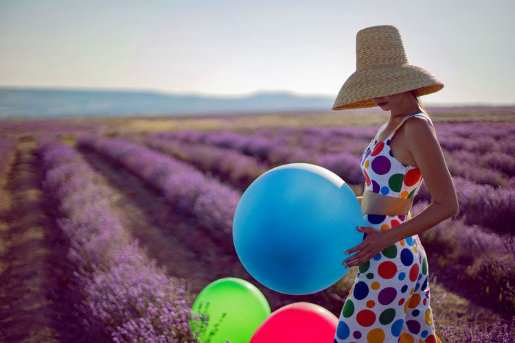 Fashionable young woman in hat on lavender field stands with colored balls and clothes to the point