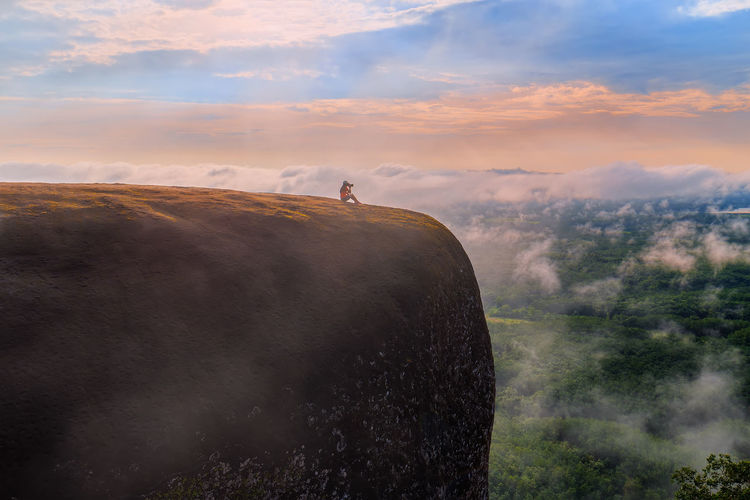 Mid distance of woman sitting on mountain against cloudy sky during sunset