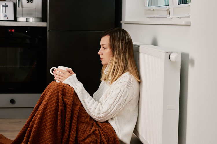 Worried woman sit near heating radiator under blanket with cup of tea
