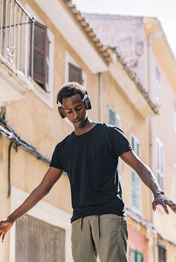 From below serious youthful african american male hipster in casual clothes and headphones listening to music in street