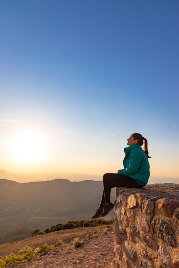 Rear view of woman sitting on rock against clear sky during sunset