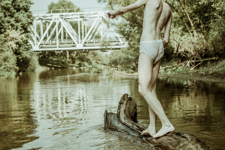 Low section of shirtless man standing on fallen tree in river at forest