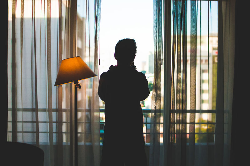 Rear view of man standing while looking through window at home