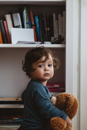 Portrait of baby girl holding teddy bear standing by shelf at home