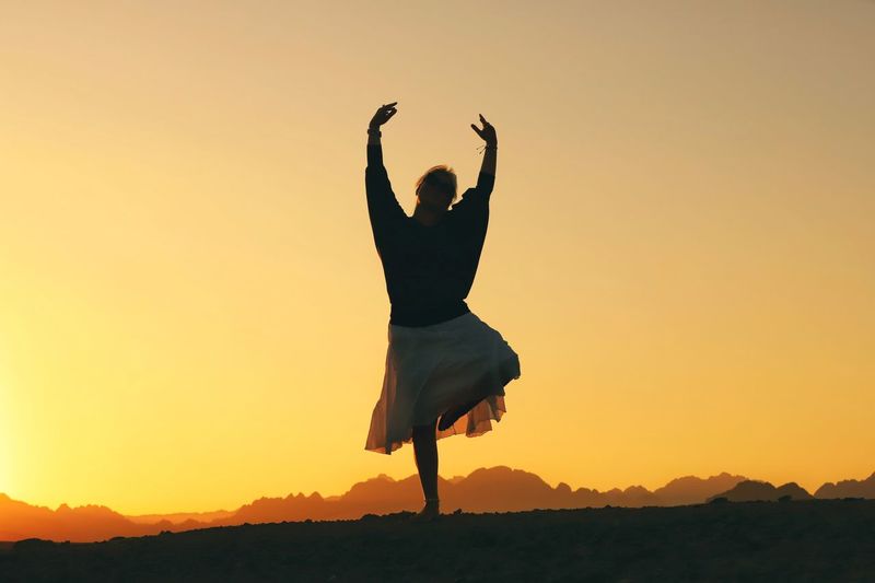 Full length of silhouette woman dancing on land against clear orange sky
