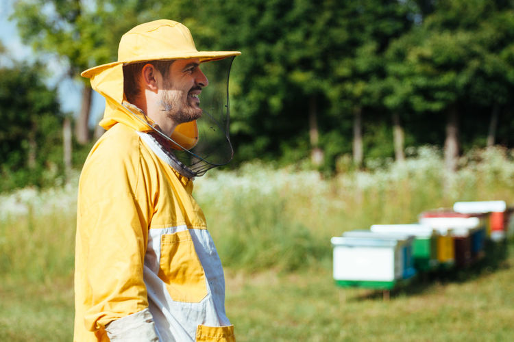 Side view of smiling beekeeper standing by beehives on land