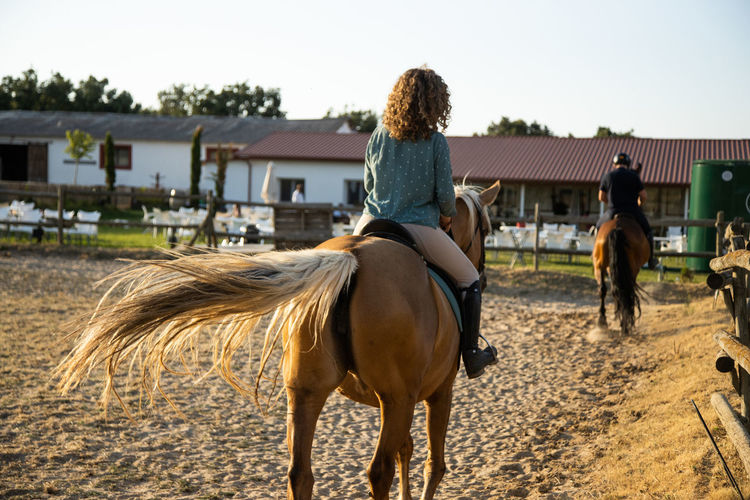 Back view of anonymous female with male trainer riding stallions in countryside on sunny day