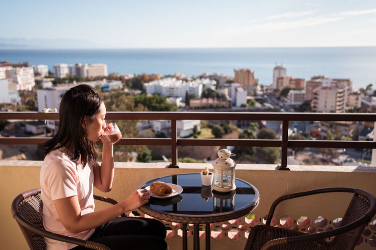 Happy asian female with glass of hot drink in hand looking at camera while sitting on balcony at table with food during breakfast